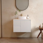 A modern bathroom scene featuring a white floating vanity cabinet with a white vessel sink, a round black-framed mirror, a small wooden side table, and a freestanding white bathtub. The room has textured beige plaster walls and a matching beige tiled floor.
