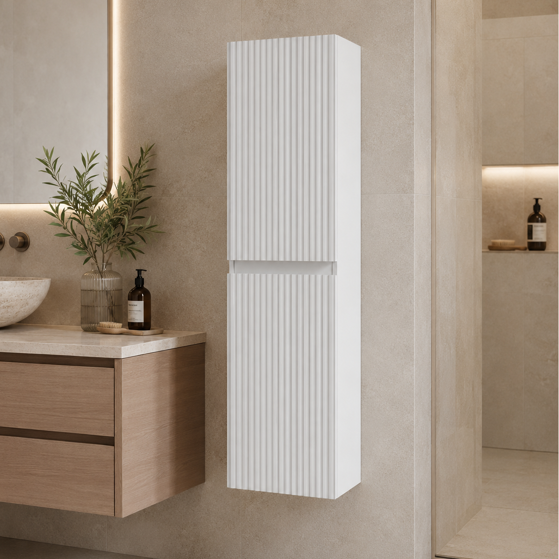 A tall, narrow white wall-mounted cabinet with vertical fluted detailing, shown in a modern beige-toned bathroom next to a floating vanity and plant.